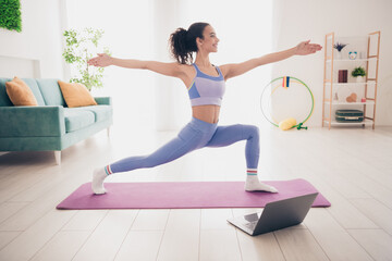 Young woman practicing yoga at home in modern interior, enjoying fitness session with laptop guidance on purple mat