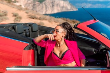 A woman in a pink suit is sitting in a red convertible. She is wearing sunglasses and a necklace.