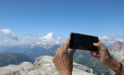 hand holds a smartphone capturing photos and videos of the Dolomite mountain peaks in the European Alps of Northern Italy during the summer