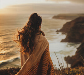 Fototapeta premium Young woman wrapped in a cozy wool blanket standing on the edge of a cliff looking at the ocean