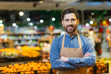 Confident male grocery worker wearing apron stands with crossed arms in front of fresh produce...