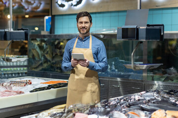 Male fishmonger in apron at seafood counter. Smiling with tablet in hand, showcasing technology use...
