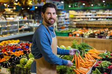 Grocery worker arranges fresh carrots in produce section. Smiling, wearing apron. Surrounding shelves filled with colorful vegetables and fruits. Emphasis on freshness, organic produce.