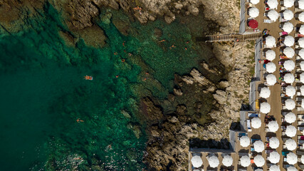Perpendicular aerial view of a beach club with white umbrellas on a rocky coast of Salento, Puglia,...
