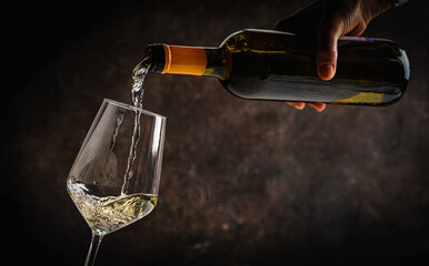 Pouring white wine into the glass on rustic dark background. Pour alcohol, winery concept.