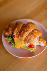 Сroissant sandwich on a pink plate, placed on a wooden table. The croissant is filled with lettuce, a slice of cheese, mayonnaise, and bright red peppers, creating a colorful and appetizing look. 