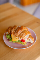 Сroissant sandwich on a pink plate, placed on a wooden table. The croissant is filled with lettuce, a slice of cheese, mayonnaise, and bright red peppers, creating a colorful and appetizing look. 