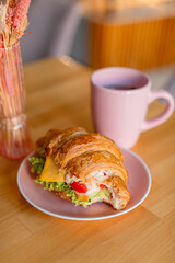 Сroissant sandwich filled with cheese, lettuce, and vegetables, served on a pink plate. Behind it, there is a matching pink cup, creating a soft and cozy atmosphere with a warm, blurred background.