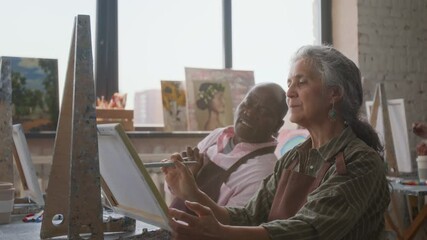 Black man looking at canvas of Caucasian senior woman and discussing picture with her while sitting next to each other at art lesson and painting