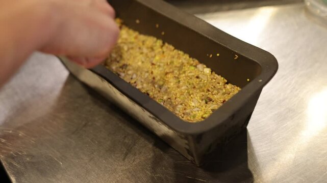 Person pressing, baking tin, grain snack. Close-up person pressing mixture seeds, nuts, grains into rectangular baking tin using spoon. Baking tin has well-used, rustic appearance, homemade dish