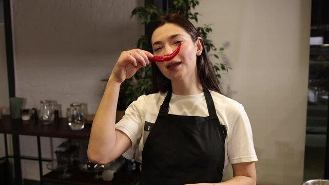 Woman apron, black apron, chili mustache. Woman wearing black apron and holding red chili pepper under her nose, mimicking mustache. Her exaggerated facial expression adds humor to scene