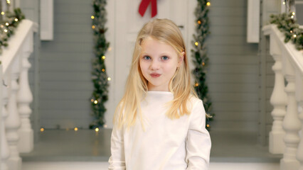 Fototapeta premium Blonde girl posing with a playful pout in a festive setting. A young blonde girl is seen playfully pouting while standing in front of a festive porch decorated with greenery and holiday lights.