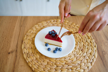 Hands hold a knife and fork in front of a plate of cheesecake against the background of the kitchen. Cheesecake on a plate. A woman eats a delicious dessert.