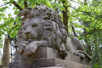 A detailed stone lion sculpture rests on a pedestal amidst lush greenery in Highgate Cemetery, London, showcasing intricate carving and a serene expression.