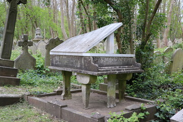 A gravestone designed as a grand piano with a raised lid, set amidst lush greenery and traditional headstones in Highgate Cemetery, London.
