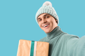 Happy young man with Christmas gift taking selfie on blue background, closeup