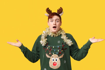 Confused young man in warm sweater, tinsel and reindeer headband on yellow background