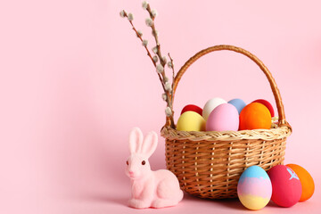 Wicker basket with painted Easter eggs, bunny and willow branch on pink background