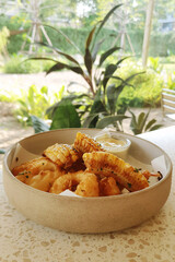Bowl of Calamari and grilled corn served with a dipping sauce, placed on a light-colored table with a garden view in the background.