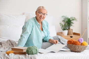 Mature woman with knitting yarn reading magazine in bedroom