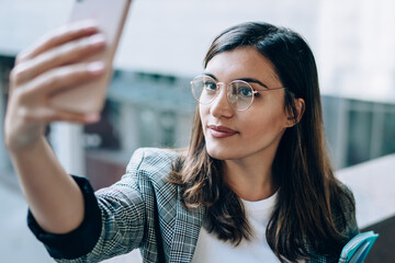 Attractive millennial hipster girl in stylish eyewear clicking selfie photos via front camera on modern cellular gadget, beautiful woman with textbook for education taking pictures via app