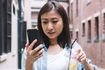 Young Asian female tourist with cold water lemon beverage in hand installing new location app for...