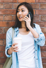 Young female person talking on mobile standing on urban brick wall background, attractive Chinese...