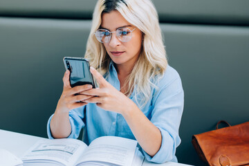 Millennial caucasian hipster girl in optical spectacles checking notification for smartphone device during break from studying with books in school environment, concept of knowledge and education