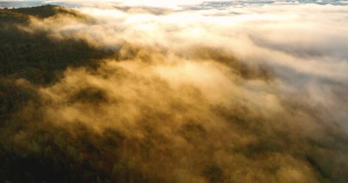 Drone aerial view descending during sunrise, with warm light bathing the clouds, mist, and fog in golden hues. The mist drifts over the colorful fall foliage, creating a serene autumn atmosphere.