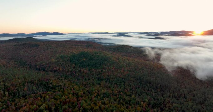 Aerial view of a mountain range covered in vibrant fall foliage, with mist and fog drifting over the colorful forested hills, capturing the beauty of the autumn season.
