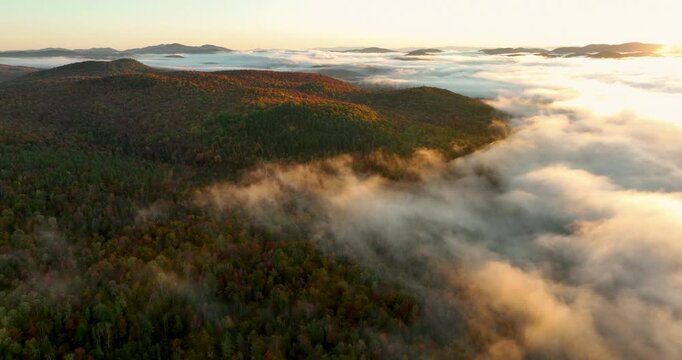 Aerial view of a mountain range covered in vibrant fall foliage, with mist and fog drifting over the colorful forested hills, capturing the beauty of the autumn season.