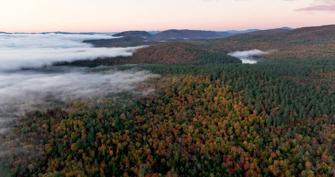 Aerial view of a mountain range covered in vibrant fall foliage, with mist and fog drifting over the colorful forested hills, capturing the beauty of the autumn season.