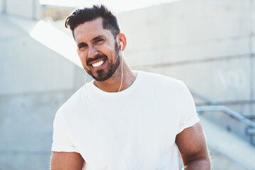 portrait of cheerful man dressed in blank t shirt enjoying free time for listening music songs via electronic headphones,happy male with modern smartphone device smiling at camera