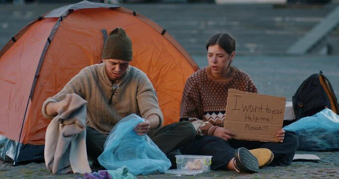 A homeless man and woman sit on the ground in front of a tent in the city. The man digs through his belongings while the woman holds a cardboard sign asking for help. Ideal for showcasing hardship.