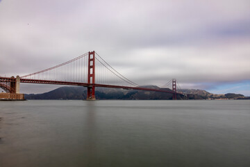 Obraz premium Serene view of the Golden Gate Bridge under a cloudy sky, capturing its iconic architecture and the tranquil waters below.