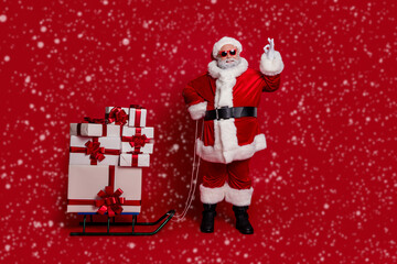 Full length photo of funky santa show okey bring gifts by sled wear cap glasses coat pants shoes isolated on red color background
