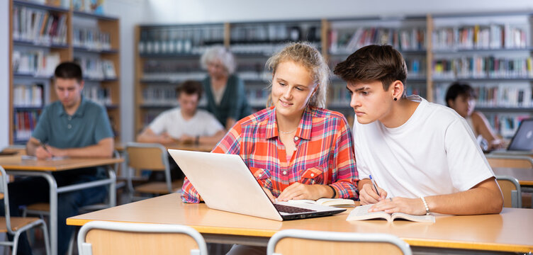 European fifteen-old-year schoolgirl, studying with a classmate in the school library on a laptop and making abstract in the copybook, preparing for lessons