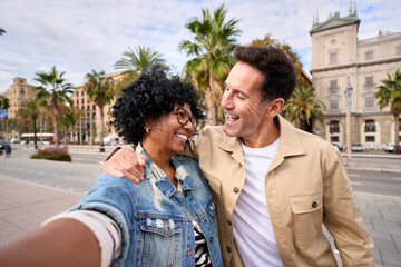 Happy multi-ethnic mature tourist couple taking selfie photo looking at each other hugged together outdoor. Middle-aged people in love make tourism smiling enjoying holidays in city street