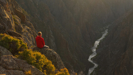 Hiker sitting on edge of cliff overlooking river inside deep canyon in Colorado, USA