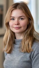 Fototapeta premium A young woman with long blonde hair looks directly at the camera with a soft smile. She is wearing a gray sweater.