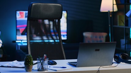 Dimly lit apartment with office desk in front of smart TV displaying news program broadcasting. Laptop on table and television screen in empty room showing channel, camera A close up shot