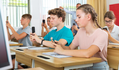 Fototapeta premium Group of modern teenagers sitting at lesson in classroom, using mobile phones..