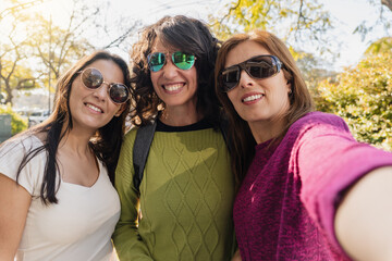 Three women wearing sunglasses smiling for a selfie in a sunny outdoor park