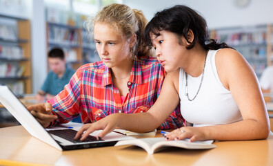 Obraz premium Two fifteen-year-old schoolgirls studying on a laptop and writing abstract in copybooks, doing homework in the school library