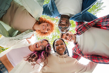 Low angle view of a group of multiracial happy young teenagers hugging each others in a circle, looking down at camera with smiling faces, representing friendship, diversity, and togetherness outdoors