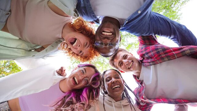 Low angle view of a group of multiracial happy young teenagers hugging each others in a circle, looking down at camera with smiling faces, representing friendship, diversity, and togetherness outdoors