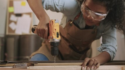 Professional African American female carpenter worker wearing apron standing using electric power drill and screwdriver to assemble by screws to fix into the wooden at workplace in furniture factory. 