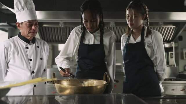 Professional Asian senior male chef in uniform with chef hat teaching cooking methods to female multiracial students listening intently and are ready to learn practical actions in restaurant kitchen.