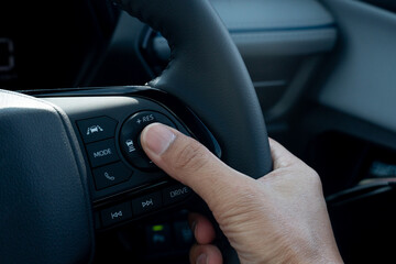 Hand of driver pressing a button on a car steering wheel. Steering wheel is black and has a leather or rubber grip. Hand is position in a way suggests the driver is using their thumb to press button.