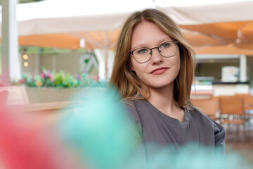 fair-haired teenage girl sits quietly at a table in a summer cafe, her expression thoughtful and...